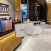 A white phone case with a gold trim and a magnetic charging ring is shown hanging in front of a cafe counter. Náhled obrázku produktu