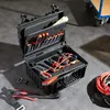 An open tool case with various tools inside, placed on a concrete floor with some cables and a cart in the background. Voir les images du produit