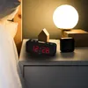 A digital alarm clock is placed on a nightstand next to a bed, with a lamp and other decorative items in the background. Produktbild-Vorschau