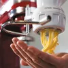 A hand is holding a KitchenAid pasta maker attachment while fresh pasta is being extruded into the hand. Náhled obrázku produktu