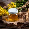A small glass jar of honey sits on a burlap coaster on a wooden surface, surrounded by yellow flowers and dried grasses. Podgląd zdjęcia produktu