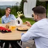 A couple enjoying a meal outdoors at a round wooden table with watermelon, cookies, and drinks. Produktbild-Vorschau