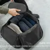 A person is packing a black duffel bag with clothes and shoes. A small black pouch is being placed into the bag. Produktbild-Vorschau