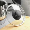 A close-up of a glass pot lid with a black handle, resting on a wooden cutting board next to a stainless steel pot. Podgląd zdjęcia produktu