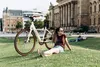 A woman sits on the grass next to a white e-bike, with a large building in the background. Another person is lying on the grass in the background. Produktbild-Vorschau