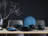 A table setting with blue and black stoneware dishes, a black vase with branches, and chairs in the background. Náhľad obrázku produktu
