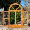 A rusty metal garden decoration shaped like an arched window with open shutters, placed on a stone wall in a garden setting. Produktbild-Vorschau
