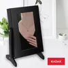 A black jewelry box with a mirror is displayed on a white surface, reflecting a person wearing a necklace. A plant and artwork are visible in the background. Produktbild-Vorschau
