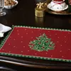 A Christmas-themed placemat with a decorated Christmas tree is laid out on a dark wooden table, with other festive items like a gold box and a teacup in the background. Produktbild-Vorschau