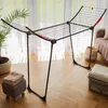 A black foldable drying rack is set up in a room with large windows and sheer curtains. There is a potted plant to the left, a small stool with towels on it, and a laundry basket with folded towels to the right. A sofa is visible in the background. Produktbild-Vorschau