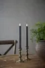 Two LED candles in brass candleholders on a wooden table with a plant in a pot and a chair in the background. Produktbild-Vorschau