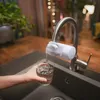 A person is filling a glass with filtered water from a faucet attachment in a kitchen sink. Produktbild-Vorschau