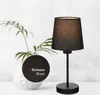 A black table lamp with a fabric shade is displayed on a white surface, with a plant in a white vase in the background. A circular swatch showing the fabric color is overlaid on the image. Produktbild-Vorschau