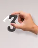 A hand is holding a black number '3' from a wall clock kit. Náhled obrázku produktu