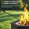 A black fire pit with a poker tool in use, set in a grassy outdoor area with trees in the background. Náhled obrázku produktu