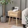 A wooden nightstand with a rattan drawer front is placed next to a sofa and a large potted plant. Náhled obrázku produktu