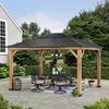 A wooden gazebo with a black roof is set up on a paved patio, furnished with a dining table and chairs. It is surrounded by lush green trees and bushes, with a house visible on the left. Produktbild-Vorschau
