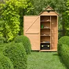 A wooden garden shed with its door open, revealing shelves with gardening tools and a toolbox, set in a lush green garden with manicured bushes and trees. Produktbild-Vorschau