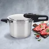 A stainless steel pressure cooker is shown on a light gray textured surface, with raw meat and herbs on a wooden cutting board in the background. Podgląd zdjęcia produktu