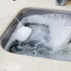 A white, bulb-shaped device with a wire is submerged in a sink filled with soapy water, suggesting it is being used to clean dishes. Podgląd zdjęcia produktu
