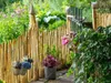 A rustic garden fence made of wooden stakes, with hanging flower pots and lush greenery. Produktbild-Vorschau