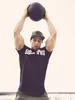 A man is holding a medicine ball above his head, wearing a black t-shirt and a green cap. Náhľad obrázku produktu
