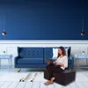 A woman sits on a dark gray folding mattress in a modern living room, working on a laptop. The room features a blue sofa, blue walls, and white trim. Náhled obrázku produktu