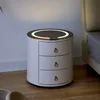 A white, cylindrical nightstand with three drawers and a round top featuring a glowing ring and wireless charging symbol, placed on a wooden floor next to a bed. Voir les images du produit