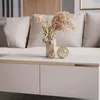 A close-up view of a modern living room coffee table with decorative items and a sectional sofa in the background. Náhľad obrázku produktu