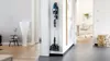 A cordless vacuum cleaner is mounted on a wall in a hallway, with a doorway leading to an office space in the background. Produktbild-Vorschau