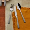 A set of cutlery, including a fork, knife, and spoon, laid out on a textured surface with an orange napkin. Produktbild-Vorschau