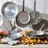 A collection of metal cookware, including frying pans and a griddle, arranged on a wooden rack. Some raw ingredients like meat and potatoes are also visible. Náhľad obrázku produktu