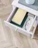 A white nightstand with an open drawer containing books, glasses, and a small box, resting on a light wood floor. Náhled obrázku produktu