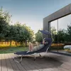 A woman relaxing on a blue chaise lounge with a canopy on a wooden deck, with trees and a modern house in the background. Náhled obrázku produktu