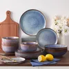 A collection of blue and gray stoneware dishes and bowls are arranged on a countertop, with a wooden cutting board and a vase of white flowers in the background. Náhled obrázku produktu