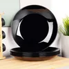 A stack of black plates on a wooden surface, with a blurred background of fabric and a plant. Podgląd zdjęcia produktu