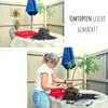 A woman is repotting a plant on a table outdoors, with a blue umbrella in the background. The top part of the image shows the plant with its roots exposed, and text overlay. Produktbild-Vorschau