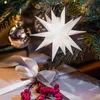 A white star ornament hangs on a Christmas tree, with a wrapped gift and red berries in the foreground. Produktbild-Vorschau