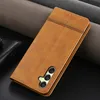 A brown leather phone case with a camera cutout is shown on a dark background with textured gray fabric rolls behind it. Produktbild-Vorschau