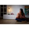 A woman sits on the floor in a living room, holding a mug and looking away from the camera. A lamp and decorative items are on a white cabinet behind her. Produktbild-Vorschau