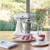 A white KitchenAid stand mixer is on a wooden table, with macarons, raspberries, and sugar in bowls nearby. A vase with flowers is in the background, with a blurred outdoor scene visible through a window. Produktbild-Vorschau