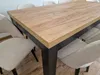 A close-up view of a wooden dining table with black legs and beige chairs around it. Produktbild-Vorschau