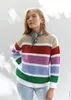 A woman wearing a striped crochet sweater and jeans, with dappled light from trees in the background. Produktbild-Vorschau