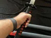 A hand holding the handle of a katana with a black and red grip, with two sheathed swords in the background. Voir les images du produit