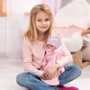 A young girl is sitting on the floor, holding a baby doll. The doll is dressed in pink and wearing a hat, and has a pacifier in its mouth. The girl is wearing a pink long-sleeved shirt and blue jeans. The background is a softly lit room with pink accents. Produktbild-Vorschau