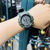 A person's arm wearing a digital watch, with a blurred background of a fence and some yellow objects. Voir les images du produit
