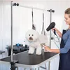 A woman is grooming a white dog on a grooming table. The dog is secured by straps, and the woman is using a hairdryer and a brush. Produktbild-Vorschau