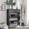 A shoe cabinet with a drawer and two flip-down compartments, filled with shoes, is placed in a hallway with striped wallpaper. A lamp, a vase with plants, and framed pictures are on top of the cabinet. A coat is hanging on a hook on the wall. Náhľad obrázku produktu