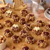 A collection of chocolate and nut-covered cookies arranged on a wooden surface, with a coffee maker and a wooden rocking horse in the background. Produktbild-Vorschau