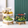 Two multi-tiered fruit stands are displayed on a white surface against a green, ribbed background. The stand on the left is made of bamboo and has white ceramic bowls filled with fruit. The stand on the right is also bamboo with dark green bowls filled wit Náhled obrázku produktu